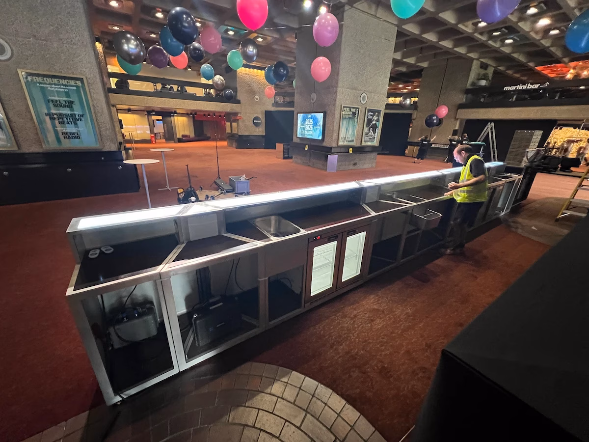 Event crew assembling a modular mobile bar setup with integrated under-counter fridges inside a venue hall.